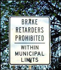 An informational street sign (white background with black letters) declaring brake retarders (engine compression brakes) are prohibited within municipal limits.