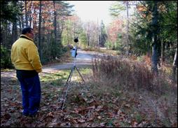 A noise analyst performs sound level measurements with a sound level meter in a forested area adjacent to a low volume road.