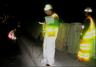 A noise analyst, wearing appropriate safety gear, uses a sound level meter to capture an individual nighttime construction operation.