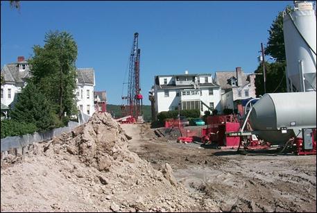 A construction area adjacent to nearby residences has several pieces of stationary equipment, such as a crane and mixing container, under operation.