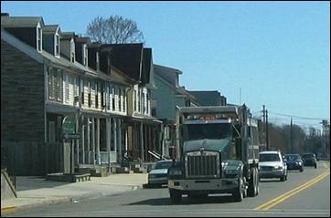 An individual dump truck in existing automobile traffic in an urban setting with nearby townhomes.
