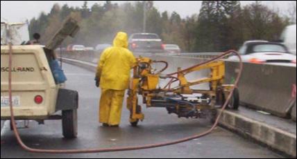 A worker operates a compressor and bridge cleaning equipment while traffic passes by in the background. 
