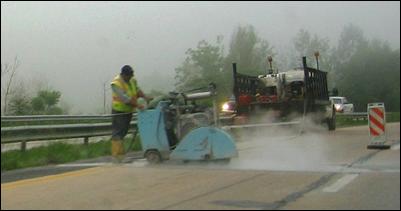A worker operates a concrete saw perpendicular to travel direction with an idling truck in the background.