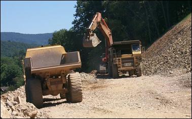 A dump truck awaits rocks from a loader while another dump truck waits in line; rock excavation is a noisy activity.