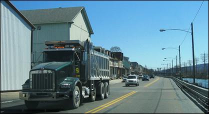 A dump truck hauling excess material from a construction site transports its cargo on a local roadway with automobile traffic present and residents adjacent to the roadway.
