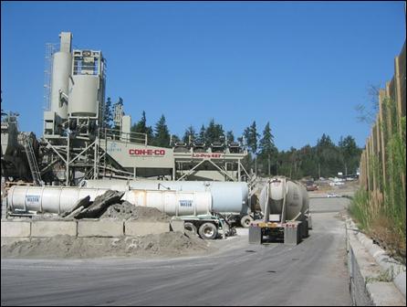 A concrete batch plant operation uses several pieces of stationary equipment along with an arriving cement mixing truck; a temporary wooden noise barrier blocks the operation noise for a community to the right.