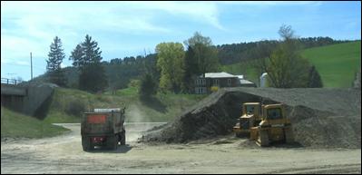 Several bulldozers form a stockpile of fill material while an unloaded dump truck exits the construction site. The fill material is placed between the construction equipment and a residence.