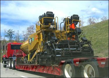 A paver is transported to a construction site on a heavy truck with an oversized trailer.