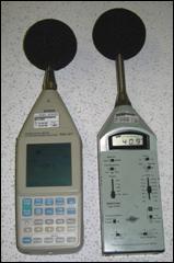 Two Type I sound level meters are placed side-by-side for comparison. Both have a spherical windscreen protecting the microphone and both have many functions available on their touchpad.