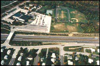An aerial photo showing an existing 6-lane highway and overpass adjacent a community of homes with a nearby recreational area with a track and sports fields.