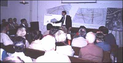 A speaker presents a project at a public meeting with many citizens in attendance.