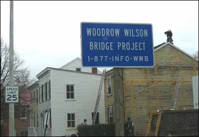 A blue sign posted on a residential street defining the site as the Woodrow Wilson Bridge Project and a telephone number to contact for information, 1-877-INFO-WWB.