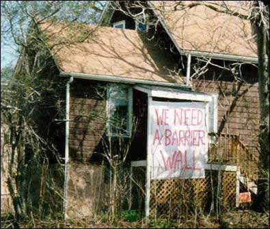 A homemade painted sign posted on a residence declaring the resident’s desire for a noise barrier. The sign reads: “We need a barrier wall”.