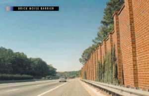 photo:  brick noise barrier, constructed immediately adjacent to the shoulder of a highway, protected by a w-beam guardrail and with some vegetation beginning to grow up the face of the barrier (photo is intended to illustrate a typical example of a well-designed and well-constructed brick noise barrier)