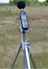 Title: Sound Level Meter - Description: A sound level meter attached to a tripod angled toward the roadway from a field with tall grasses.