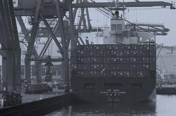 Photo. Loading dock with freight containers, ship, and trucks.