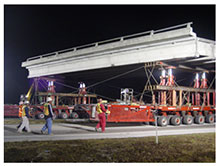 Self-propelled modular transporters (SPMTs) carry a bridge span down a road. The operator of the computer that controls the SPMT movement is walking behind the SPMTs, along with three other construction workers monitoring progress.