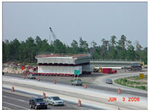 A new bridge span supported by self-propelled modular transporters is shown in a staging area where the span was constructed near the bridge location.