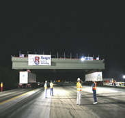 Self-propelled modular transporters carry a bridge span up a road to the bridge site during nighttime construction. Four construction workers with safety vests monitor progress.