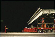 Self-propelled modular transporters (SPMTs) carry a bridge span down a road. The operator of the computer that controls the SPMT movement is walking behind the SPMTs.