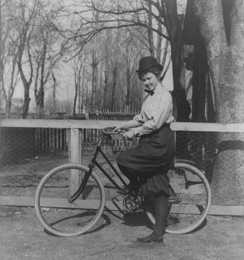 Black and white photograph showing a young woman posing next to a bicycle.