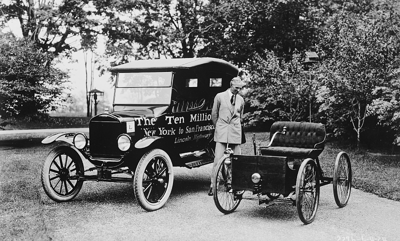 Black and white photograph of a gentleman standing between the first and the ten millionth Ford automobiles.