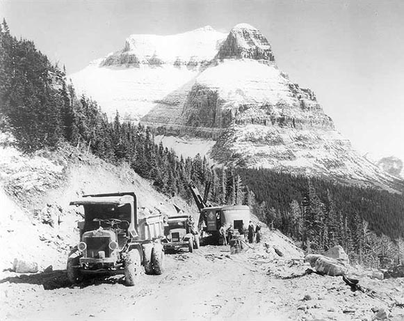 Black and white photograph of a machinery at a construction site with the Going‐to‐the‐Sun Mountain in the background.