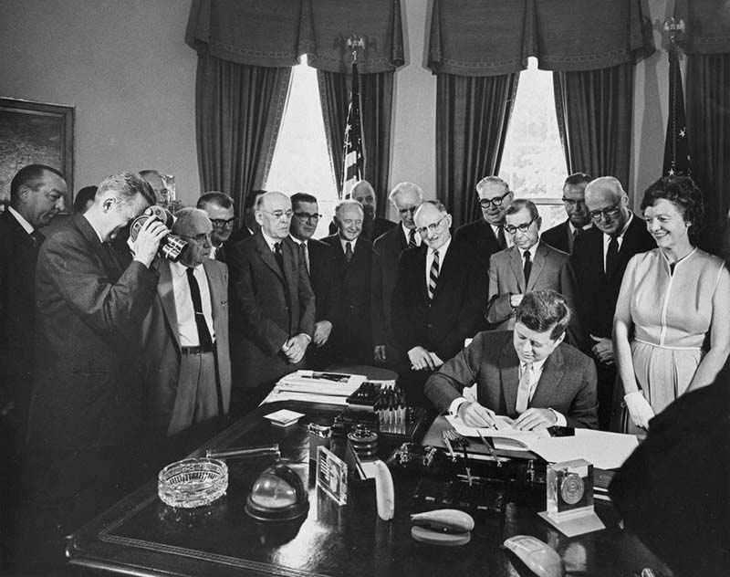 Black and white commemorative photograph of President Kennedy signing the Federal Highway Act in the Oval Office.