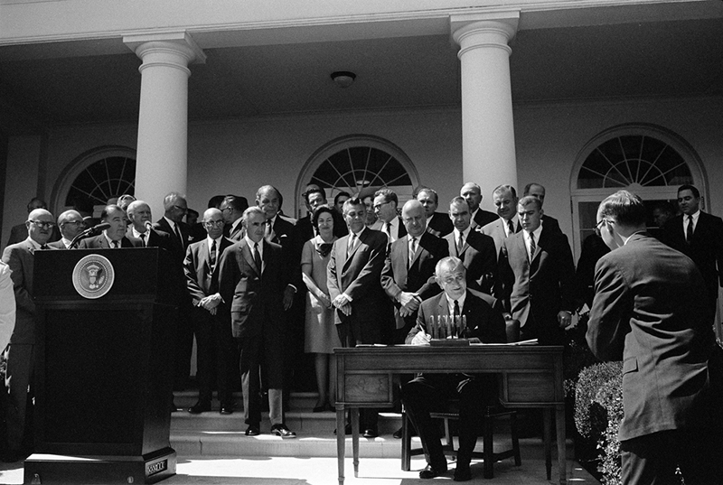 Black and white commemorative photograph of President Johnson signing the National Traffic and Motor Vehicle Safety Act in the Oval Office.