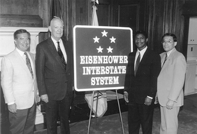 Photograph of John Eisenhower, son of President Eisenhower, with the Eisenhower Interstate System sign.