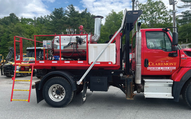A red municipal flatbed truck equipped with a white tank, hook-lift body, and side ladder parked outdoors. 