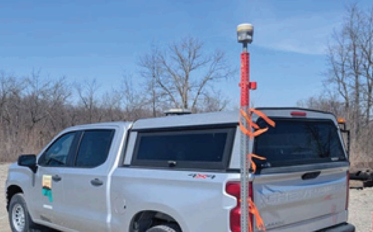 A pole-like device is attached to the rear of a white pick-up truck with a camera mounted at the top. 