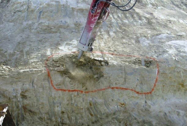 Photo. A hydraulic hammer sculpting a rock face (the material to be removed has been outlined with common marking
                paint).