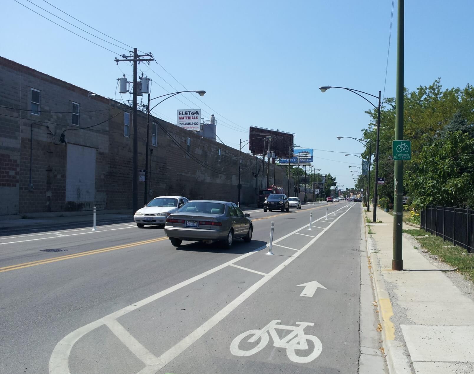 Vehicles are shown on a two-lane, two-way city street. There is a marked buffered bike lane on each side, and pylons provide further delineation between vehicles and the bike lane.