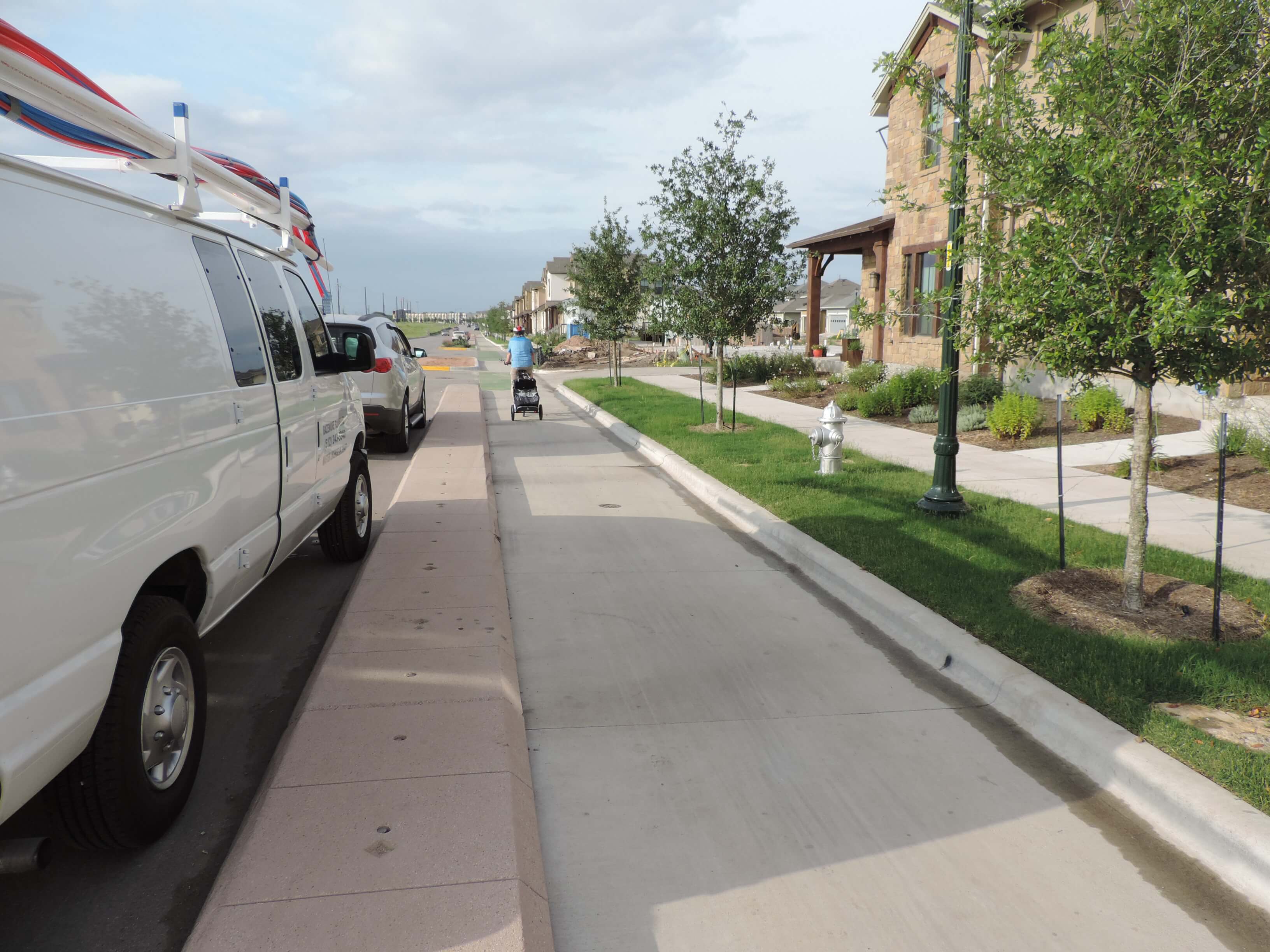 Parked vehicles are shown in a residential area, with a wide curb between the parked vehicles and the bike lane. A person wearing a helmet is pedaling a bicycle with a trailer in the bike lane. A grassy strip separates the bike lane from the sidewalk.