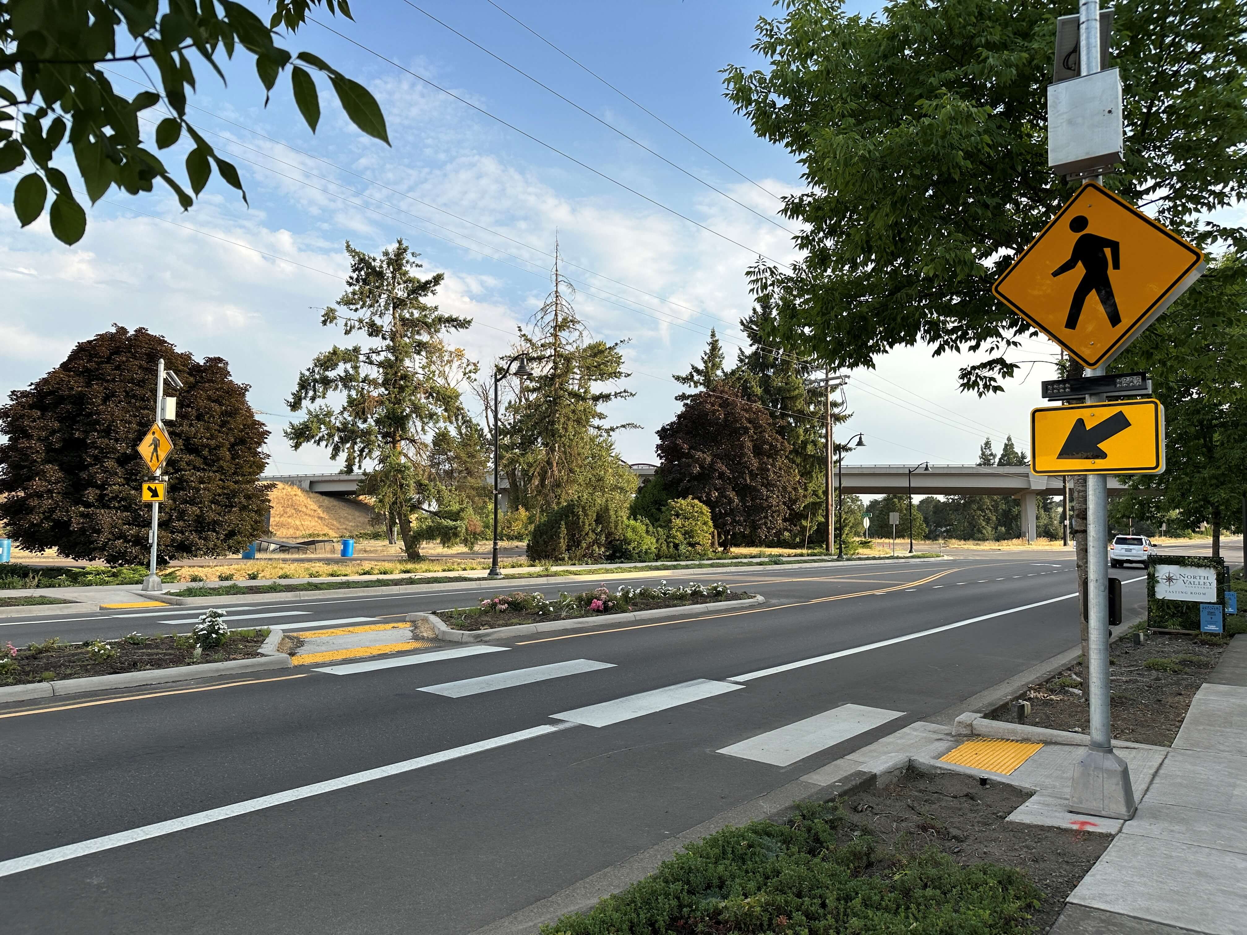 A four-lane road includes a crosswalk that has a rectangular rapid flashing beacon at the pedestrian crossing, pedestrian crossing signs, and curb ramps on both sides, clear pavement markings throughout, and a refuge island.