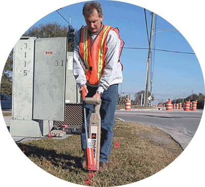 This picture depicts 'designating' and shows an operator interpreting the presence of a subsurface communications line using a pipe and cable locator and marking its horizontal position with orange paint on the ground using a paint dispenser.