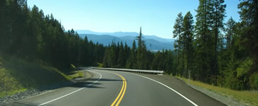 Curvilinear downgrade with guardrail surrounded by trees with mountains in the background.