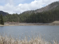 Photo of the completed bridge from the far side of Fenton Lake. The bridge is nearly hidden from view at the base of the pine-covered mountains across the valley.