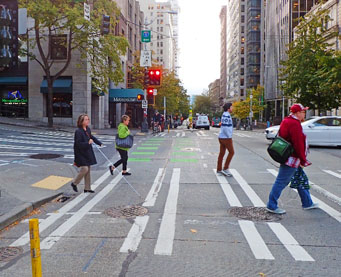Photo of people crossing in the crosswalk on a busy downtown street. On of the people has a vision disability and is using a cane.