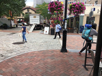 Photo of a shared street intersection. The surface of the comfort zone and crosswalk are smooth brick. The surface of the shared zone is cobled. Pedestrians are walking in various directions across the photo, including in the shared zone.
