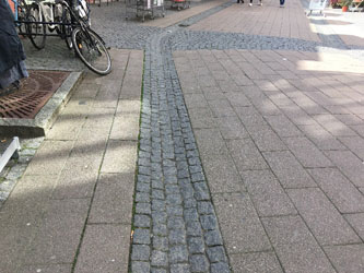 Photo of the edge of a valley gutter at the edge of a shared street. The vally gutter is made of small granite cobbles which contrast in color and texture with the adjoining pavers.