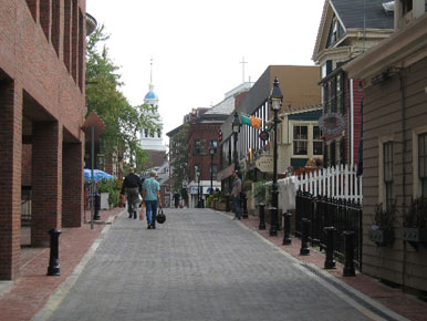 Photo of a shared street that has limited right-of-way. There is a strip of red brick at either side that might be interpreted as a comfort zone, but these spaces are not wide enough to be accessible. No pedestrians can be seen in these spaces. All of the pedestrians in the photo are walking in the shared zone.
