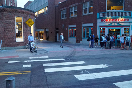 Photo of a location where a shared alley intersects a conventional street. There is a detectable warning surface at the end of the alley placed in advance of a driveway apron that leads to the intersecting street, which a man is walking directly toward from the shared alley. This detectable warning is not aligned with a marked crrosswalk. Instead, a marked crosswalk is shown to the left. where a man with a stroller is waiting to cross.
