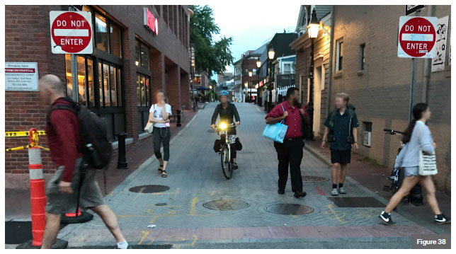 Photo of a the gateway of a shared alley. Several pedestrians and a bicyclist are seen exiting the street. Two pedestrians are seen crossing.