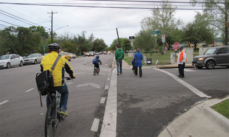 Photograph of bicyclists (in biycle lane) and pedestrians crossing an intersection at a marked crossing  with a crossing guard directing traffic.