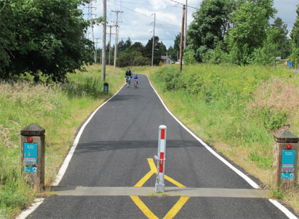 Photo of a person riding a bicycle on a path. There are bollards at the front end of the path. The center bollard is removable.