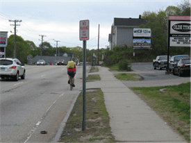 Image shows a bicyclist navigating a busy road with many driveways and no dedicated bicycle facility.