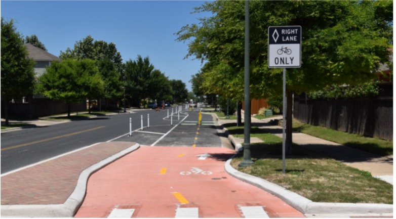 This image shows a two-way protected bicycle lane. The forefront of the image shows a crosswalk at the start of the lane. The lane is painted light pink in the crosswalk area. To the left of the bike lane is a two-lane road. There are trees on each side of the road. To the right of the bike lane is a sign that says 'Right Lane Only' with a bicycle icon.