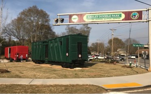This image shows two railcars, one red and one green, sitting in a park. There is a large sign that says 'Easley Doodle Park.' There is grass and a sidewalk in the foreground and trees, parked cars, a street, and sky in the background. There are a few people walking around.